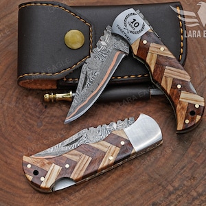 May include: Two folding knives with patterned wooden handles and Damascus steel blades are displayed with a brown leather case. One knife has the text "10 YEARS TOGETHER" on the blade. The knives are on a wooden surface.