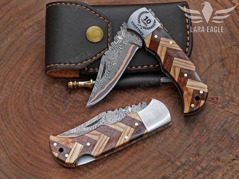 May include: Two folding knives with patterned wooden handles and Damascus steel blades are displayed with a brown leather case. One knife has the text "10 YEARS TOGETHER" on the blade. The knives are on a wooden surface.