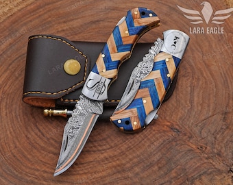 May include: Two folding knives with blue, brown, and light wood chevron patterned handles. The blades have a decorative pattern. One knife is open, and the other is closed. A brown leather sheath is behind the knives.