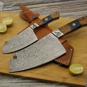 May include: Two cleavers with wooden handles and personalized engravings, "A" and "S Smith", on a wooden cutting board with lemons. The cleavers have a Damascus steel blade with a hammered finish.
