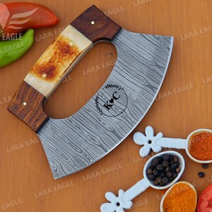 Op de afbeelding: Een gepersonaliseerde, gebogen, roestvrijstalen cleaver met een houten en beenderen handvat. Het blad heeft een damastpatroon en is gegraveerd met de initialen "K & C".