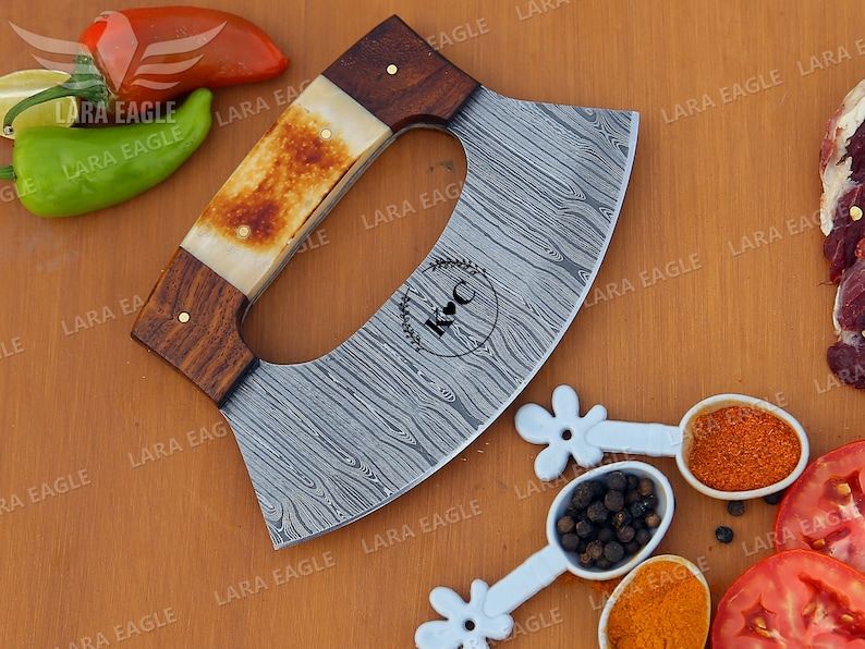 Op de afbeelding: Een gepersonaliseerde, gebogen, roestvrijstalen cleaver met een houten en beenderen handvat. Het blad heeft een damastpatroon en is gegraveerd met de initialen "K & C".