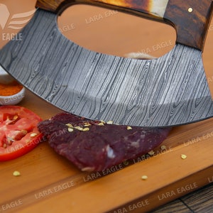 Op de afbeelding: Een close-up van een gebogen, roestvrijstalen mes met een houten handvat. Het mes ligt op een houten snijplank met een stuk rauw vlees en een tomatenplak.