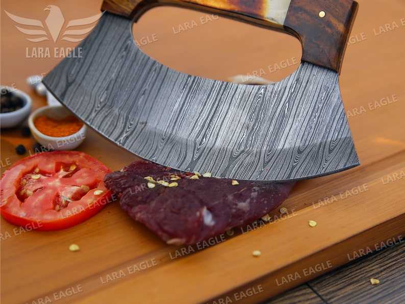 Op de afbeelding: Een close-up van een gebogen, roestvrijstalen mes met een houten handvat. Het mes ligt op een houten snijplank met een stuk rauw vlees en een tomatenplak.