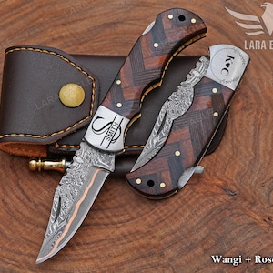 May include: Two folding knives with patterned blades and wooden handles, one open and one closed, next to a brown leather sheath. The handles have a dark wood grain pattern with brass accents. The open knife has the name "SMITH" on the blade.