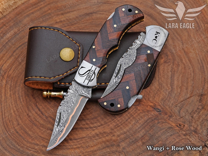 May include: Two folding knives with patterned blades and wooden handles, one open and one closed, next to a brown leather sheath. The handles have a dark wood grain pattern with brass accents. The open knife has the name "SMITH" on the blade.