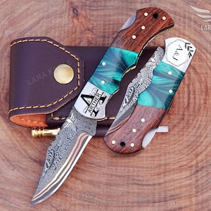 May include: A folding pocket knife with a Damascus steel blade and a handle featuring wood and green resin. The knife is engraved with the name "ARTHER" and initials "A&J". A brown leather sheath is also included.
