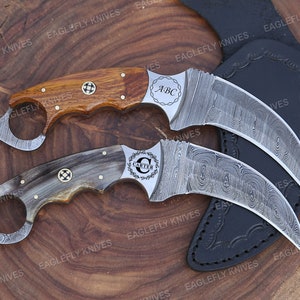 May include: Two personalized karambit knives with Damascus steel blades. The first knife has a wooden handle with the initials "ABC" engraved on the blade. The second knife has a horn handle with the name "CARTER" engraved on the blade. Both knives are displayed on a black leather sheath.