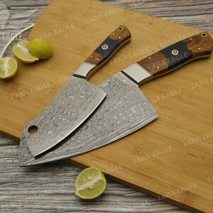 May include: Two Damascus steel cleavers with wooden handles on a wooden cutting board. The cleavers have a hammered finish and a decorative pattern on the blades. The handles are made of dark wood with a blue accent.