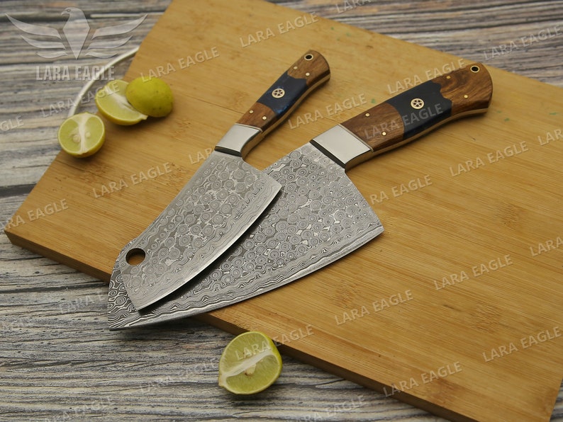 May include: Two Damascus steel cleavers with wooden handles on a wooden cutting board. The cleavers have a hammered finish and a decorative pattern on the blades. The handles are made of dark wood with a blue accent.