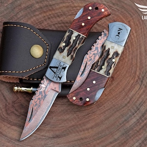 May include: Two folding knives with wooden handles and patterned blades, one open and one closed, next to a brown leather sheath. The open knife has the name "ARTHER" engraved on the blade. The closed knife has the initials "K♡C" engraved on the blade.