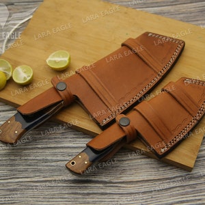 May include: Two leather sheaths for knives, one with a curved blade and one with a straight blade. The sheaths are brown leather with decorative stitching and are lying on a wooden cutting board.