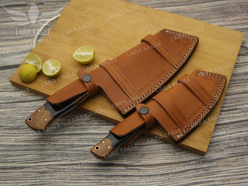 May include: Two leather sheaths for knives, one with a curved blade and one with a straight blade. The sheaths are brown leather with decorative stitching and are lying on a wooden cutting board.