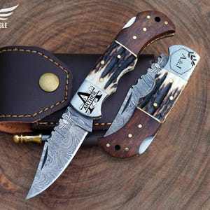 May include: Two folding knives with ornate blades and wooden handles, one open and one closed, rest on a wooden surface. The open knife displays the name "ARTHER" and the closed knife has the initials "A&J". A brown leather sheath is also present.