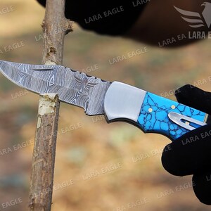 May include: A folding pocket knife with a blue and white patterned handle. The blade is made of Damascus steel and has a decorative eagle etched into the metal.