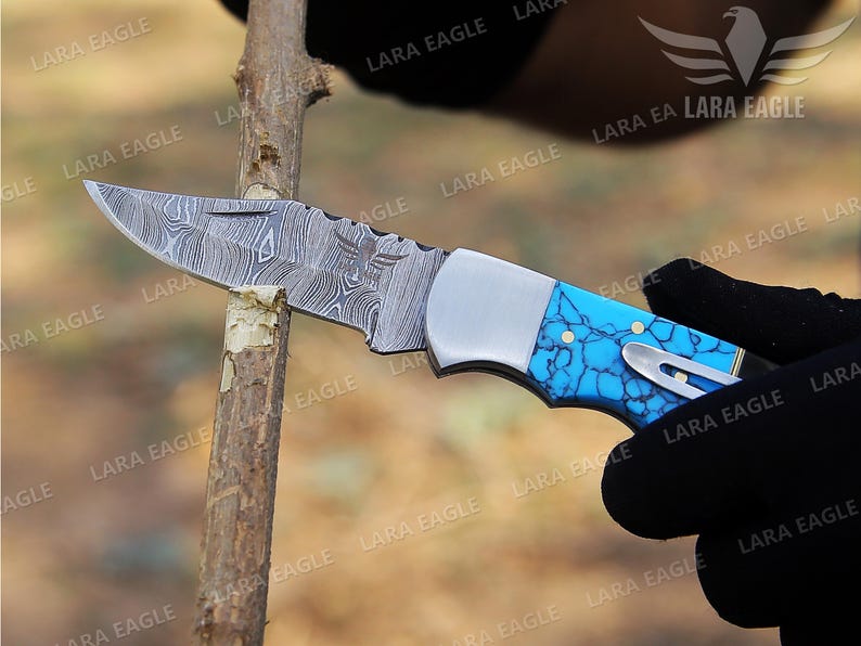May include: A folding pocket knife with a blue and white patterned handle. The blade is made of Damascus steel and has a decorative eagle etched into the metal.