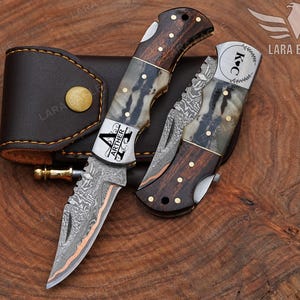 May include: Two folding knives with wooden handles and decorative metal blades, one open and one closed, next to a brown leather case. The open knife has the name "ARTHER" on the blade. The closed knife has the initials "K C" on the blade.