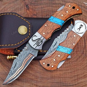 May include: Two folding knives with wooden handles, turquoise accents, and patterned blades are displayed. One knife is open, revealing the blade, while the other is closed. A leather sheath with a gold button is in the background. The knives have the name "SMITH" and "K C" engraved.