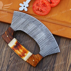 Op de afbeelding: Een gebogen, roestvrijstalen mes met een houten handvat en een decoratief patroon. Het mes ligt op een houten snijplank met tomaten.