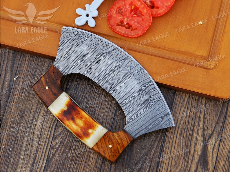 Op de afbeelding: Een gebogen, roestvrijstalen mes met een houten handvat en een decoratief patroon. Het mes ligt op een houten snijplank met tomaten.