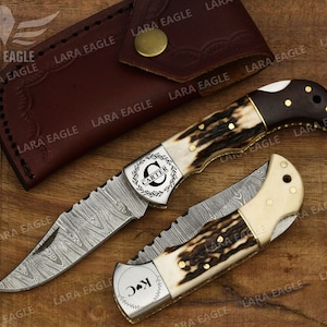 May include: Two personalized pocket knives with a Damascus steel blade and a stag horn handle. The knives have a gold-colored metal accent and are engraved with the names "Carter" and "J.C."