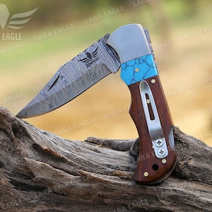 May include: A folding pocket knife with a patterned steel blade and a wooden handle. The handle has a turquoise inlay and a silver clip. The knife is open and resting on a piece of wood.