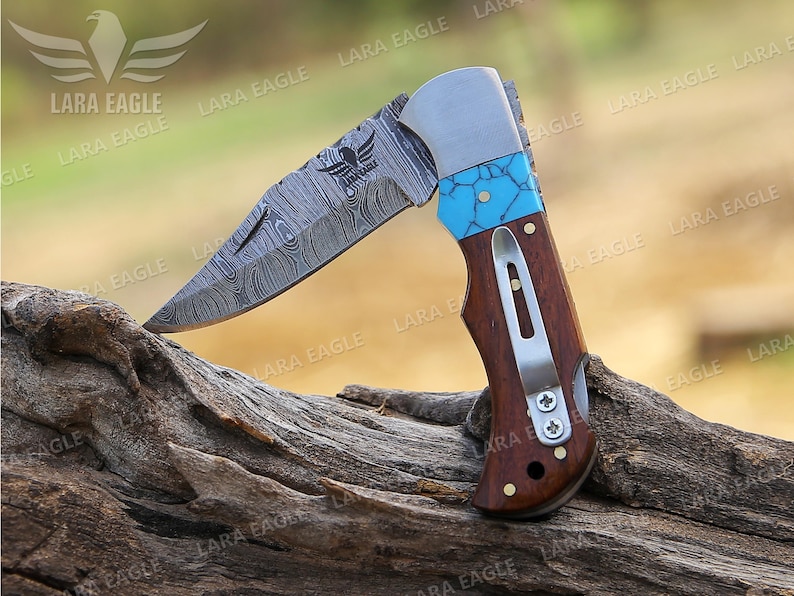 May include: A folding pocket knife with a patterned steel blade and a wooden handle. The handle has a turquoise inlay and a silver clip. The knife is open and resting on a piece of wood.