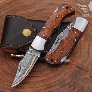 May include: Two folding knives with wooden handles and decorative blades are displayed next to a brown leather sheath. The blades have a unique pattern, and the handles are adorned with brass rivets. The knives are on a wooden surface.