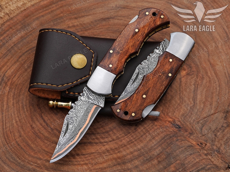 May include: Two folding knives with wooden handles and decorative blades are displayed next to a brown leather sheath. The blades have a unique pattern, and the handles are adorned with brass rivets. The knives are on a wooden surface.