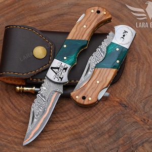 May include: Two folding pocket knives with Damascus steel blades, wood and teal resin handles, and brass pins. One knife is open, displaying the name ARTHER, while the other is closed and marked with the letters KC. A brown leather sheath is included.