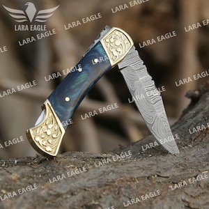 May include: A folding pocket knife with a patterned steel blade and a handle featuring a blue and gold design. The handle has a gold-coloured metal bolster with an ornate floral pattern. The knife is open, displaying the blade.