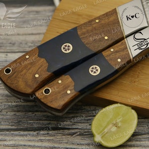 May include: Two personalized kitchen knives with wooden handles and blue resin accents. The blades are Damascus steel with a hammered finish. The handles are engraved with the initials "K&C" and "S SMITH".
