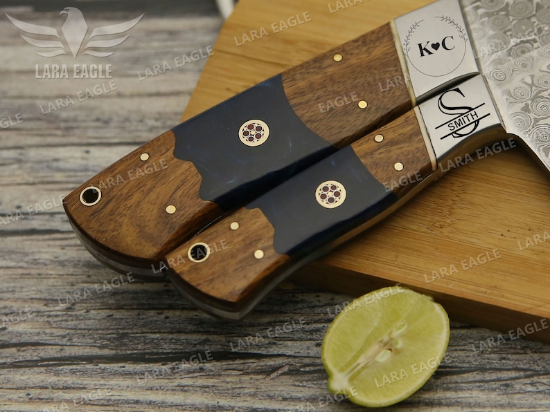 May include: Two personalized kitchen knives with wooden handles and blue resin accents. The blades are Damascus steel with a hammered finish. The handles are engraved with the initials "K&C" and "S SMITH".