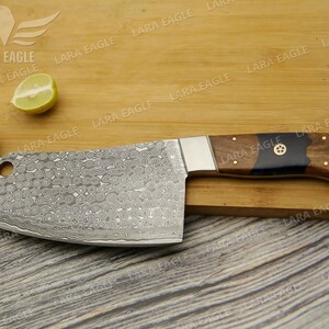 May include: A large, silver cleaver with a wooden handle. The blade has a hammered finish and a small hole near the handle. The handle is made of dark wood with a black resin inlay. The cleaver is resting on a wooden cutting board.