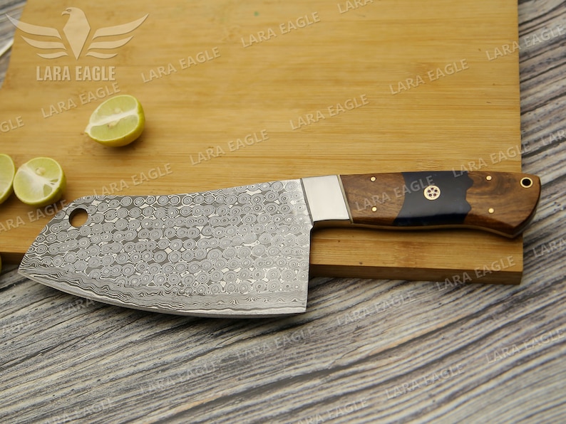 May include: A large, silver cleaver with a wooden handle. The blade has a hammered finish and a small hole near the handle. The handle is made of dark wood with a black resin inlay. The cleaver is resting on a wooden cutting board.