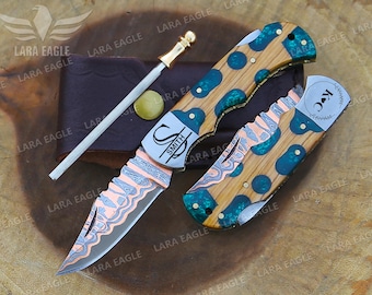 May include: A set of custom knives with wooden handles, copper accents, and a sharpening steel. The blades feature intricate patterns and the handles have teal-colored inlays. One knife is open, displaying the blade, and the other is closed.