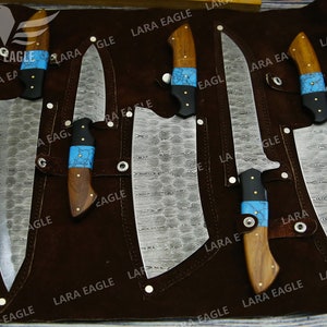 May include: Set of six kitchen knives with wooden handles and blue and black accents. The knives are displayed on a brown leather roll with the text "LARA EAGLE" repeated throughout.