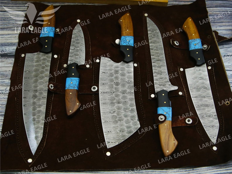 May include: Set of six kitchen knives with wooden handles and blue and black accents. The knives are displayed on a brown leather roll with the text "LARA EAGLE" repeated throughout.