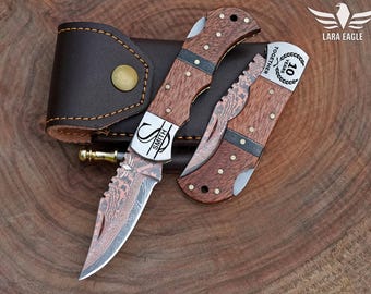 May include: Two folding knives with wooden handles and patterned blades are displayed next to a brown leather sheath. One knife is open, revealing the blade. The blades have a unique pattern. The knife has the word "SMITH" on the blade.