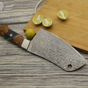 May include: A large, silver cleaver with a wooden handle. The cleaver has a patterned blade and a small hole near the tip. The cleaver is lying on a wooden cutting board with lemons on the board.
