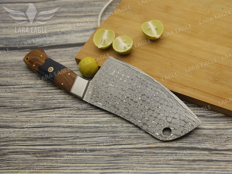 May include: A large, silver cleaver with a wooden handle. The cleaver has a patterned blade and a small hole near the tip. The cleaver is lying on a wooden cutting board with lemons on the board.