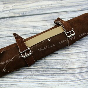 May include: Brown leather roll with two silver buckles and a tan leather strap. The roll is on a light wood surface.