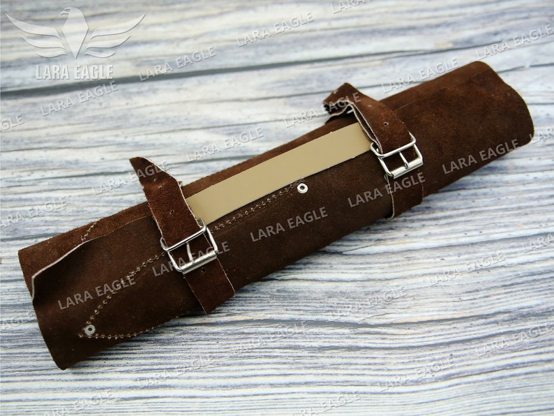 May include: Brown leather roll with two silver buckles and a tan leather strap. The roll is on a light wood surface.