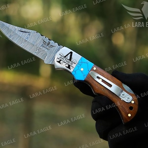 May include: A personalized pocket knife with a blue and silver handle. The blade is made of Damascus steel and has a decorative pattern. The handle is made of wood and has a metal clip. The knife is engraved with the name "Arther" and a decorative design.