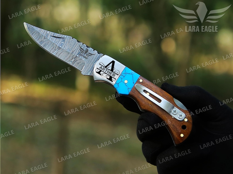May include: A personalized pocket knife with a blue and silver handle. The blade is made of Damascus steel and has a decorative pattern. The handle is made of wood and has a metal clip. The knife is engraved with the name "Arther" and a decorative design.