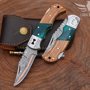 May include: Two folding knives with wooden handles and green accents, one engraved with "MICHAEL GROOMSMAN 12.10.2020" and a bow tie symbol. The other knife is engraved with "A&J". A brown leather case is also included.