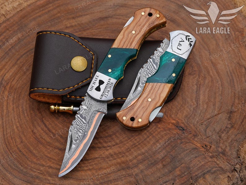 May include: Two folding knives with wooden handles and green accents, one engraved with "MICHAEL GROOMSMAN 12.10.2020" and a bow tie symbol. The other knife is engraved with "A&J". A brown leather case is also included.