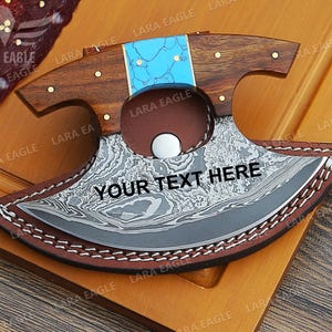 May include: A personalized, curved, stainless steel knife with a wooden handle and a turquoise inlay. The knife is in a brown leather sheath with decorative stitching. The knife is engraved with the text "YOUR TEXT HERE".