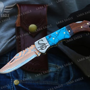 Op de afbeelding: Een gepersonaliseerd zakmes met een blauw en turquoise gepatroneerd handvat, een roestvrijstalen mes met een damastpatroon en een houten accent. Het mes is gegraveerd met de letter "S" en de naam "Smith".