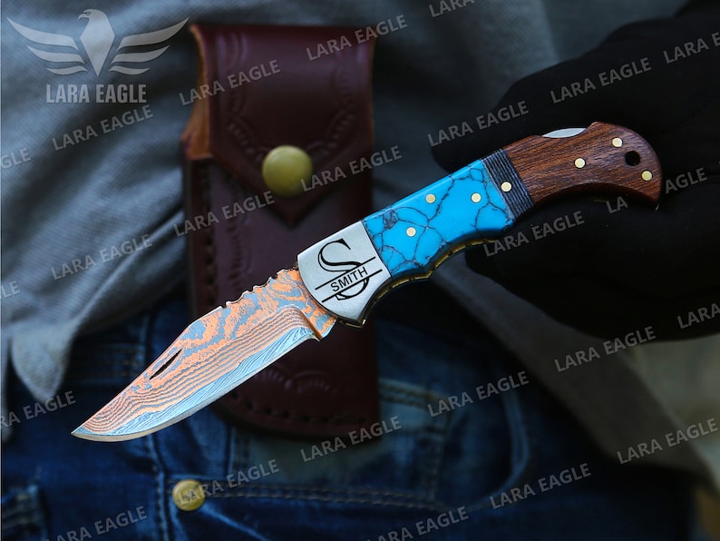 Op de afbeelding: Een gepersonaliseerd zakmes met een blauw en turquoise gepatroneerd handvat, een roestvrijstalen mes met een damastpatroon en een houten accent. Het mes is gegraveerd met de letter "S" en de naam "Smith".